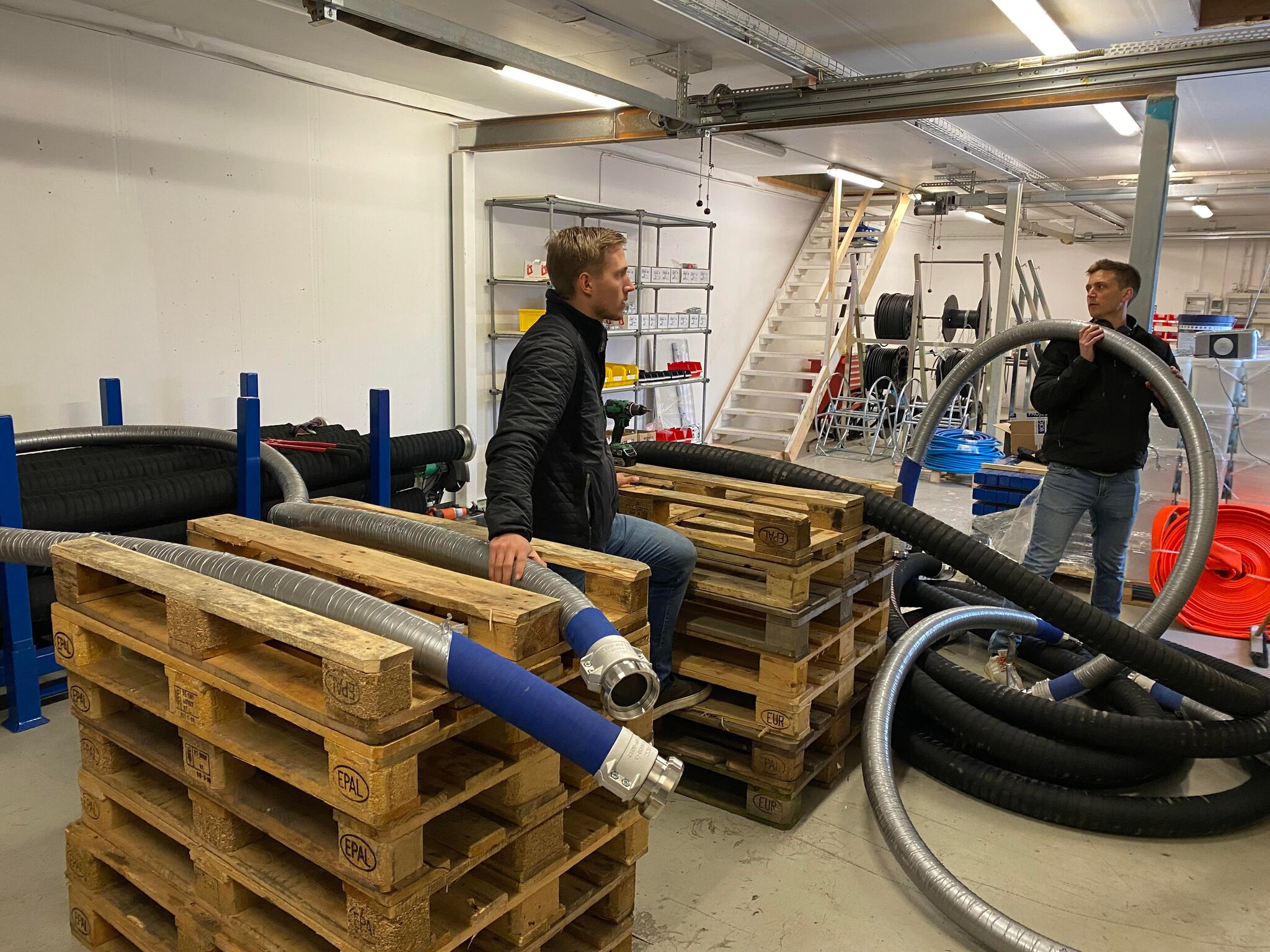 Två män som står i en lagerlokal med massa slangar och EU-pallar